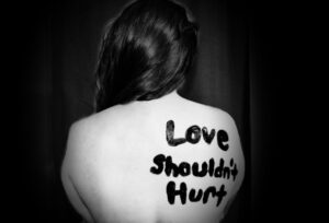 A picture of a woman sitting in a dark room with “ Love Shouldn’t Hurt” painted on her back in protest of domestic violence.