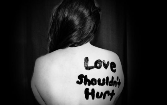 A picture of a woman sitting in a dark room with “ Love Shouldn’t Hurt” painted on her back in protest of domestic violence.